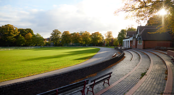 Queens Park cricket pavillion This landscape photograph features the Queen's Park cricket pavilion in Queen's Park, United Kingdom, taken during a clear autumn morning. The image shows the pavilion building, curved paths lined with benches, and a well-maintained cricket field bordered by a low brick wall. Trees displaying autumn foliage are present throughout the park, adding natural color to the scene. The sunlight casts long shadows across the grass and highlights the structure and details of the pavilion, demonstrating the harmonious blend of nature and architecture in this section of the park.
